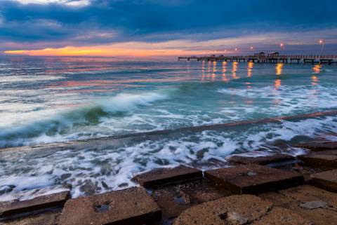 Ocean waves washing up onto stone with a pier in the background.